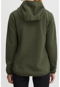 Oliwkowy polarowy hoodie o miękkiej teksturze, z kapturem i rękawami raglanowymi. Hem jest lekko zaokrąglony z tyłu. Na sobie czarne dżinsy.