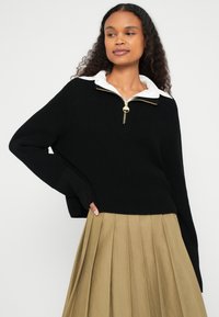 Fekete kötött pulóver cipzáros nyakkal és arany színű kiegészítőkkel, hozzá pleated khaki szoknya párosítva. A pulóver széles, bordázott mandzsettákkal rendelkezik.