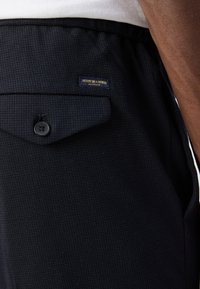 Granatowe teksturowane spodnie z widocznym guzikiem, kieszenią z patką oraz metką z napisem „Scotch & Soda, Amsterdam”. Podkreślony detal tkaniny tkanej.