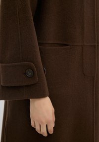 Cappotto in lana marrone con una finitura testurizzata, caratterizzato da un polsino con bottone e una tasca laterale. Il design presenta linee pulite e un minimalismo nei dettagli.