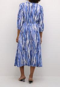 Sininen ja valkoinen kuviollinen mekko, jossa on kapeat hihat, kolmannes hihasta ja liehuva hame. Suora kangas. Korkokengät näkyvissä.