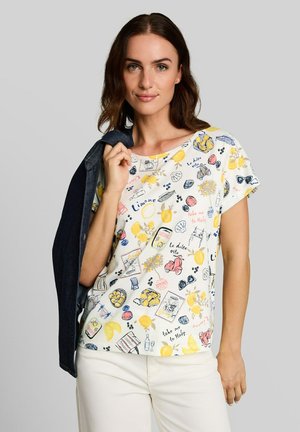 Vrouw draagt een wit bedrukt t-shirt met kleurrijke krabbels en Italiaanse zinnen, houdt een donkere jas over haar schouder tegen een effen achtergrond.