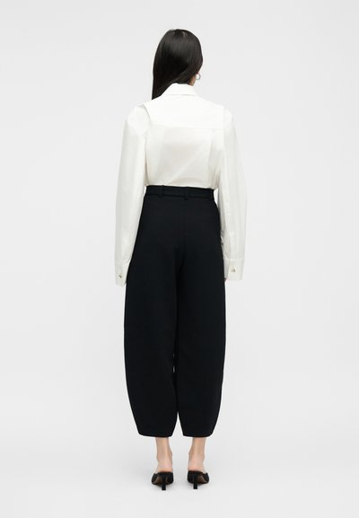 Γυναίκα με μακριά σκούρα μαλλιά φοράει λευκό μακρυμάνικο πουκάμισο περασμένο μέσα σε ψηλόμεσο, φαρδύ μαύρο παντελόνι και μαύρα παπούτσια με τακούνι, στέκεται γυρισμένη με την πλάτη.