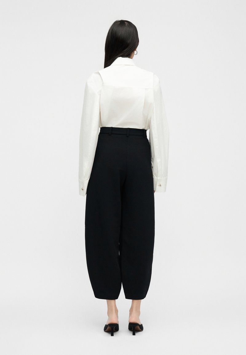 Frau mit langen dunklen Haaren trägt ein weißes langärmliges Hemd, das in locker sitzende, hoch taillierte schwarze Hosen gesteckt ist, und schwarze Absatzschuhe. Sie steht mit dem Rücken zur Kamera.