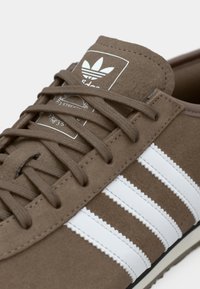 Sapatilha de camurça castanha com três riscas brancas, atacadores texturizados e língua acolchoada com uma etiqueta de logótipo na lateral.