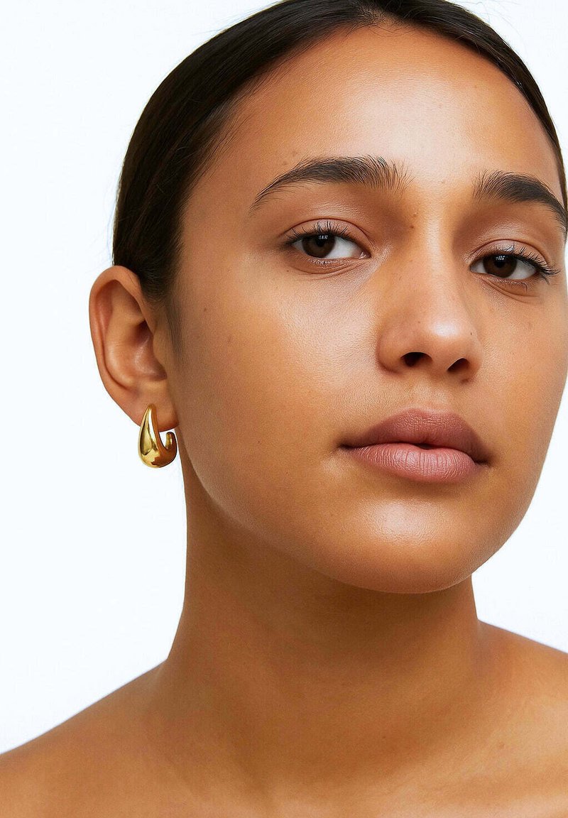 Kuldne tilgakujuline kõrvarõngas, millel on sile ja peegeldav pind, ripub lobest ja täiendab neutraalset nahatooni.