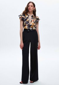 Färgglad mönstrad blus med volangärmar, kombinerad med högmidjade svarta byxor med en slät textur och knappr detaljer.