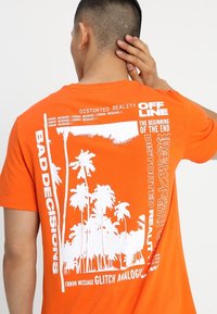 Oranžové tričko s krátkým rukávem, které má na zadní straně bílé grafické texty a design palmy, s hladkým bavlněným povrchem.