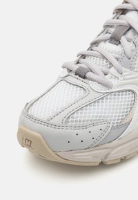 Baskets de sport gris clair en matériau mesh et cuir, avec un bout arrondi et une semelle en caoutchouc texturé. Présente des lacets gris et des accents subtils.