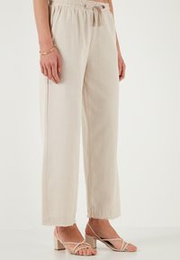 Beige, weit geschnittene Hosen aus einem leichten Stoff, mit einem elastischen Bund und einem Kordelzug sowie einem ausgefransten Saum. Kombiniert mit hellen Sandalen.