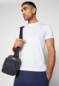 Bolso bandolera negro con textura de malla y una correa resistente, sostenido en una mano. El fondo es neutro con un degradado gris claro.