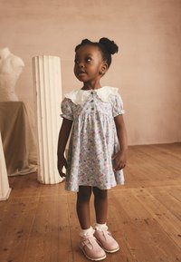 Blaues geblümtes Kleid mit kurzen Ärmeln, spitzenbesetztem Kragen und Knopfverschluss. Rosa Schuhe mit Rüschen-Socken. Hintergrund aus Holzfußboden.