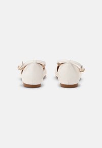 Bailarinas de cuero blanco con la parte trasera redondeada, tira al tobillo con hebilla dorada y un tacón bajo marrón. Textura suave con un diseño minimalista.
