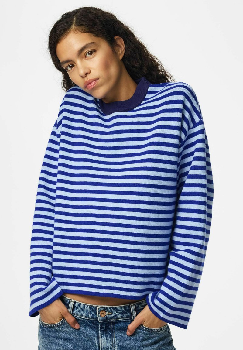 Femme aux cheveux bouclés foncés portant un pull à manches longues rayé bleu et blanc et un jean bleu, les mains dans les poches.