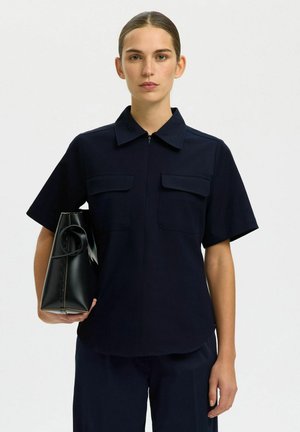 Frau trägt ein marineblaues Kurzarmhemd mit Fronttaschen und hält eine schwarze Handtasche vor einem einfarbigen weißen Hintergrund.