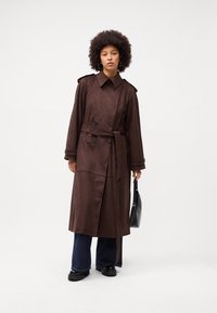 Cappotto trench in suede marrone con chiusura doppiopetto, vita con cintura e colletto tradizionale. Abbinato a jeans svasati e una borsa nera.