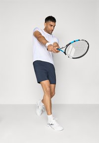 T-shirt atlético branco, calções azul-marinho e ténis brancos. Segurando uma raquete de ténis preta e azul, demonstrando um movimento de swing ativo.