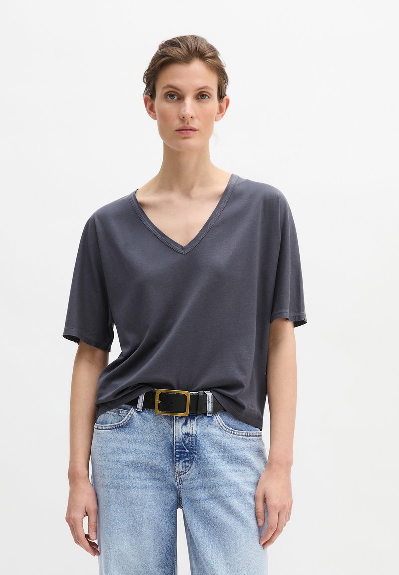 Γυναίκα που φοράει σκούρο γκρι μπλούζα με V-λαιμόκοψη και κοντά μανίκια, που έχει μπει μέσα σε ανοιχτόχρωμο τζιν παντελόνι με μαύρη ζώνη με μεγάλο χρυσό αγκράφα.