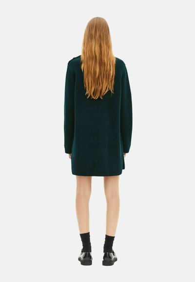 Abito in maglia verde oversize con maniche lunghe, polsini e orlo a coste, abbinato a calze nere e scarpe. La vista è da dietro.