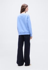 Γυναίκα που στέκεται πλευρικά φορώντας ανοιχτό μπλε φούτερ, σκούρο τζιν με φαρδιά μπατζάκια και λευκά παπούτσια σε απλό λευκό φόντο.