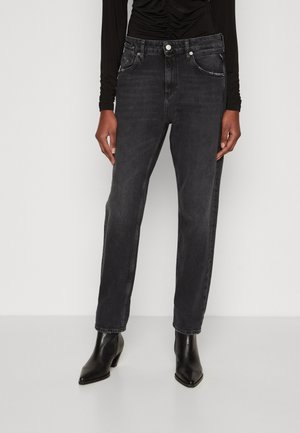 Person trägt schwarze High-Waist-Jeans mit Fronttaschen und schwarze Stiefeletten, steht vor einem einfarbigen hellen Hintergrund.