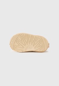 Semelle en caoutchouc beige avec un design de bande de roulement texturé. Logo visible près du talon. Profil plat avec une légère courbe aux bords des orteils et du talon.