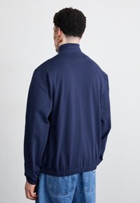 Granatowa bluza z wysokim kołnierzem, elastycznymi mankietami i ściągaczem u dołu. Miękki materiał, minimalistyczny design, widok od tyłu na jasnym tle.