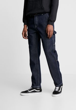 Homme portant un jean droit en denim bleu foncé avec des coutures blanches et des baskets noires à semelles et lacets blancs, debout devant un fond gris.