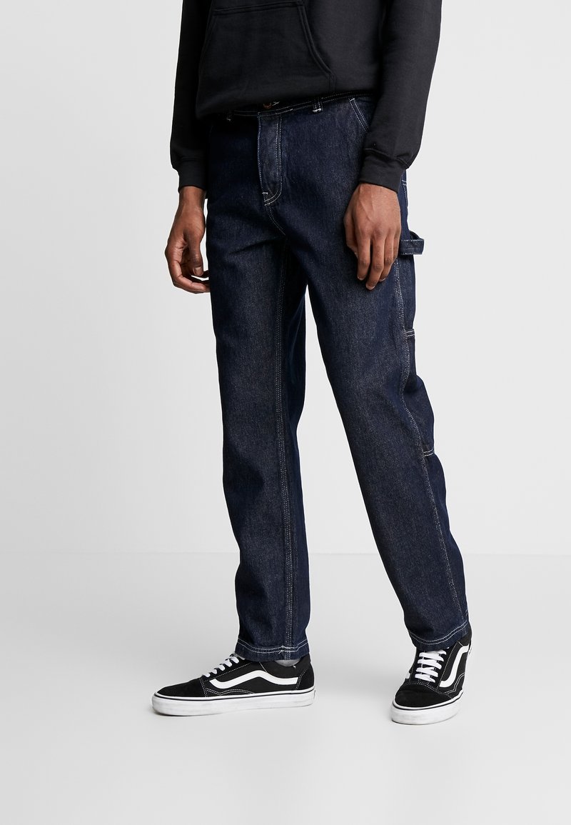 Homme portant un jean droit en denim bleu foncé avec des coutures blanches et des baskets noires à semelles et lacets blancs, debout devant un fond gris.
