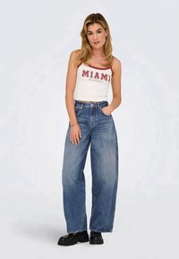 Top branco sem mangas com "MIAMI" em letras vermelhas, calças de ganga largas de cintura alta em azul, sapatos pretos chunky, modelo de pé contra um fundo claro.