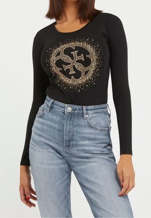 Kobieta ubrana w czarną bluzkę z długim rękawem z złotym, okrągłym wzorem krzyża oraz jasnoniebieskie, wysokowalne dżinsy, stojąca na tle białego tła.