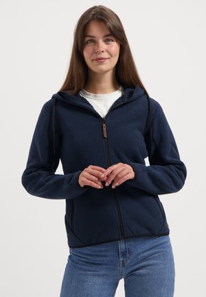 Junge Frau in einem marineblauen zip-up Hoodie mit Kapuze und blauen Jeans, die vor einem schlichten weißen Hintergrund steht.