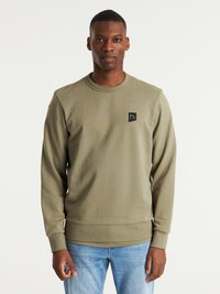 Sudadera verde oliva de tejido suave, con cuello redondo, puños acanalados y un pequeño parche de logo negro en el pecho. Se lleva con unos vaqueros.