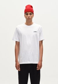 T-shirt alb cu mânecă scurtă, cu logo-ul "GABBA" negru pe pieptul stâng. Purtați cu pantaloni negri și o pălărie tricotată roșie.