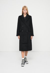 Palton negru cu dublu rând de nasturi, realizat dintr-un material moale, având un guler mare, două buzunare frontale și o croială până la genunchi.