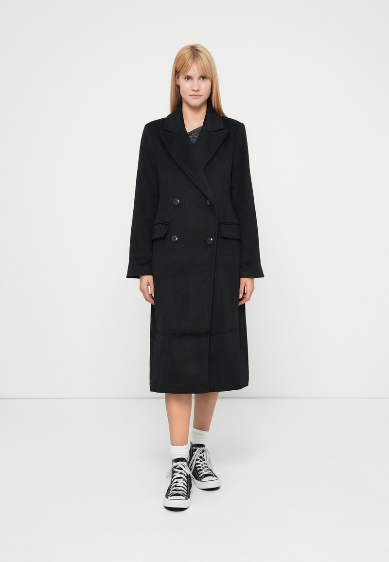 Palton negru cu dublu rând de nasturi, realizat dintr-un material moale, având un guler mare, două buzunare frontale și o croială până la genunchi.