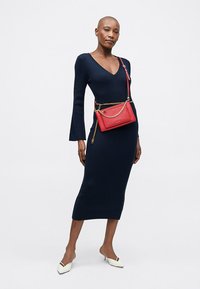 Granatowa sukienka z prążkowanej dzianiny, z długimi rękawami i dekoltem w kształcie litery V, zestawiona z czerwoną torebką na ramię z złotym łańcuszkiem. Białe czółenka w szpic.