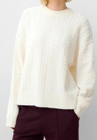 Cream cable-knit sweater with ribbed hem and cuffs. Soft texture, loose fit, featuring raised braided patterns. Paired with dark pants.