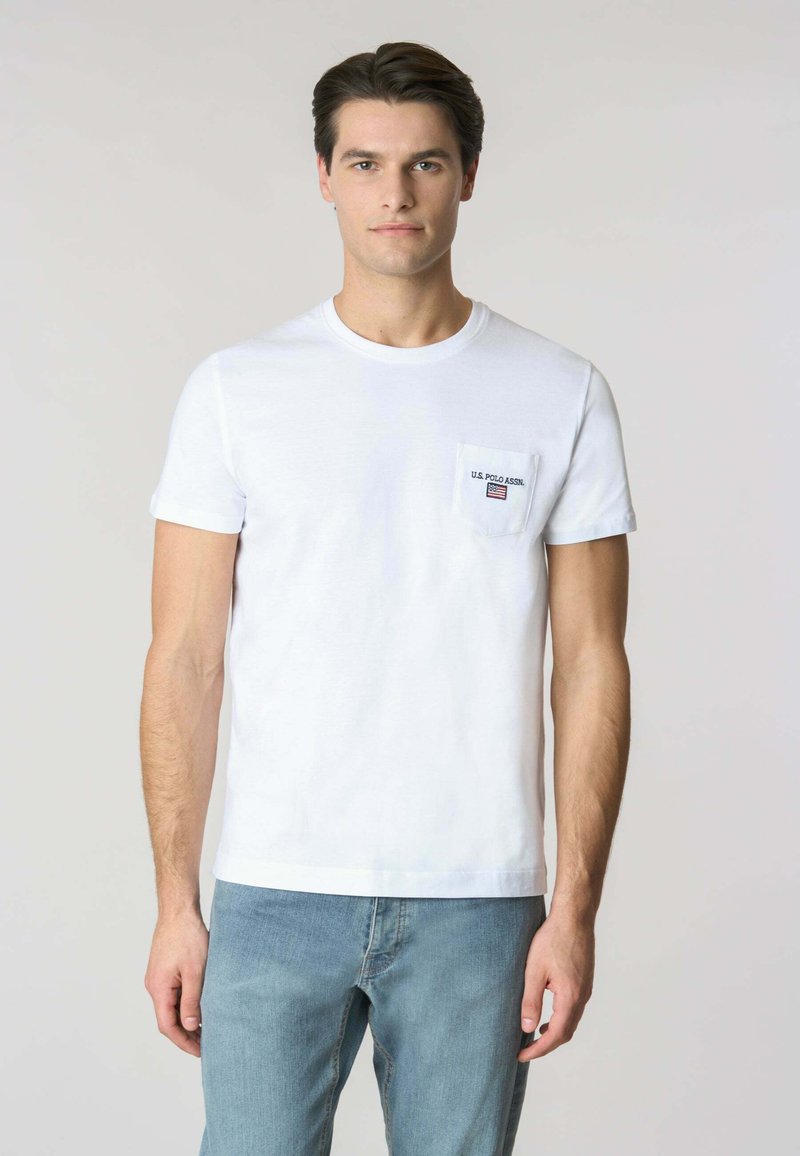 Maglietta bianca a maniche corte in cotone, con taschino sul petto e logo "U.S. Polo Assn.", abbinata a jeans azzurri chiari.