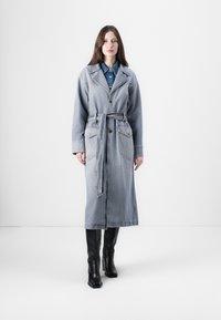 Manteau en denim bleu clair avec taille ceinturée, grandes poches avant, col à revers et manches longues. Bottes en cuir noires en dessous.