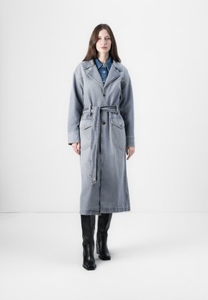 Lichte blauwe denim trenchcoat met een riem in de taille, grote voorkanten zakken, reverskraag en lange mouwen. Zwarte lederen laarzen eronder.