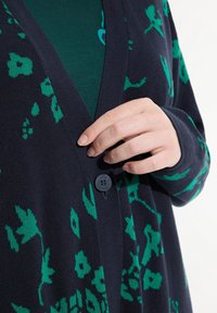 Marinblå cardigan med ett grönt blommönster, V-ringning och knappstängning. Materialet ser mjukt ut med en strukturerad yta.