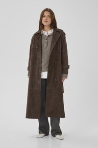 Nainen seisoo yllään pitkä ruskea kaksirivinen takki, harmaa villapaita, valkoinen paita, väljiä mustia housuja ja valkoiset kengät yksinkertaisella taustalla.