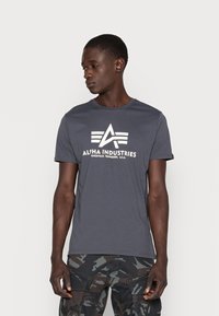 Tricou din bumbac gri cu mâneci scurte, cu un logo grafic alb "Alpha Industries" și un design unghiular. Guler plat și croială normală.