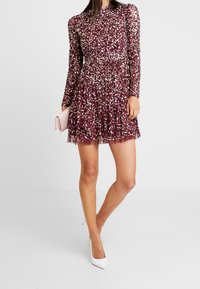 Kvinna i en vinröd långärmad paljettmini-klänning som håller en ljusrosa clutchtvätt, matchad med vita spetsiga högklackade skor.