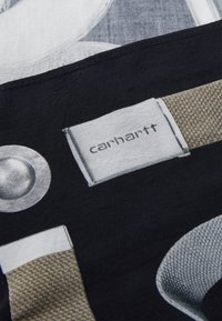 Tissu noir avec un motif imprimé comportant des éléments métalliques circulaires, des sangles texturées en beige et une étiquette portant la mention "carhartt."