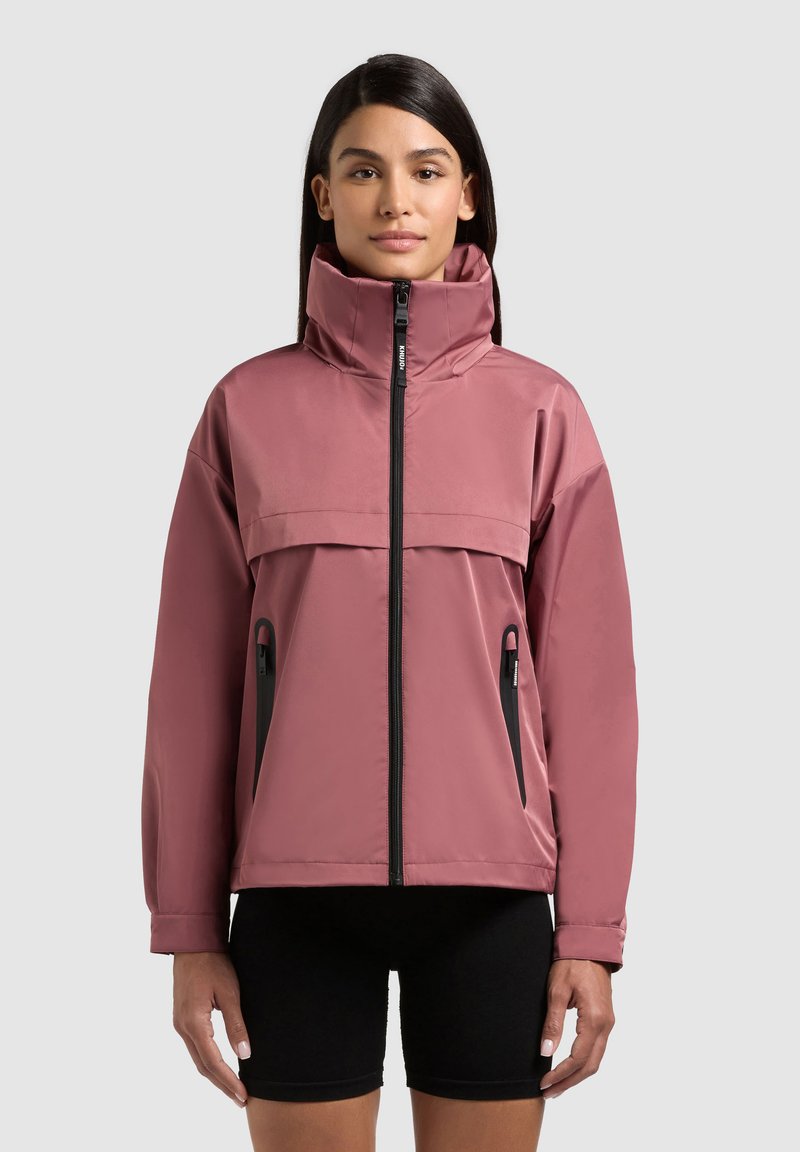 Žena oblečená v mauve mikine so zipsom a vysokým golierom so striebornými zipsovými vreckami, v kombinácii s čiernymi cyklistickými šortkami, stojaca pred svetlým pozadím.