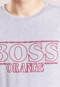 Detailní záběr na osobu v světle šedé košili s nápisem "BOSS ORANGE" tištěným velkými červenými obrysy na hrudi.