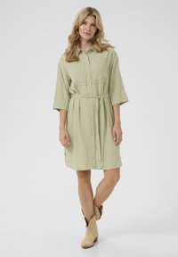 Abito camicia verde oliva chiaro con bottoni frontali, due tasche sul petto e vita con cintura. Presenta maniche a tre quarti e una texture stropicciata.