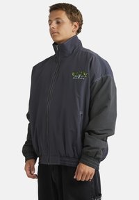 Windbreaker jacket in navy with contrasting dark gray sleeves. Features a high collar, zip closure, and embroidered logo on the chest.